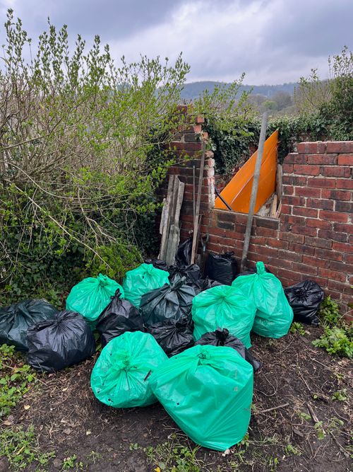 15 or more sacks of rubbish in green and black, large plastic bags. All neatly tied and arranged in a corner. Behind them larger items which would not fit including roof slates and scrap metal.