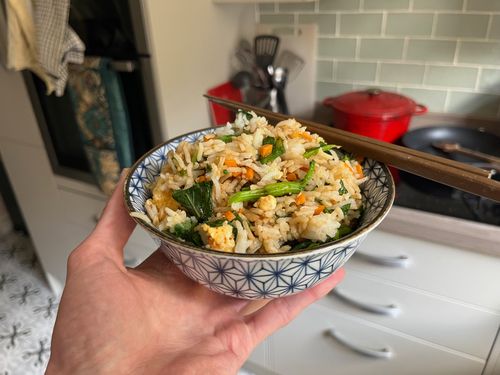 Egg fried rice with greens steams in an attractive bowl, white and decorated with a blue geometric pattern. Chop sticks rest on top.