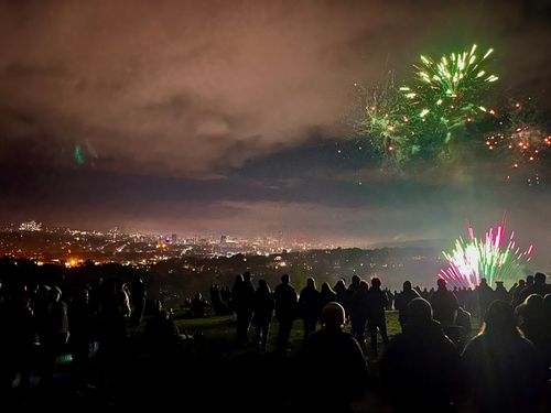 To the distance, a city aglow. Then dark, sweeping residential hills flecked with fireworks. In the foreground, a large colourful explosion illuminates a crowd.