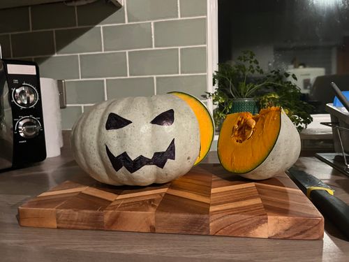 A pumpkin with Halloween-style face drew on with marker. It's friendly and menacing. It sits on a wooden chopping board, with a big wedge cut out.