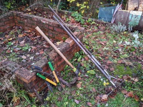 A hoe, rake, mattock, serrated blade, shears, and lopper are lent against a dilapidated cold frame.