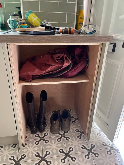 An open, plywood kitchen cabinet with an open bottom and shelf. Buddy boots sit on the floor within and backpacks have been placed onto the shelf.