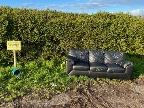 A black leather sofa sits next to a yellow warning on a post, no dumping, no tipping. It's sunny, and the sofa isn't not appealing.