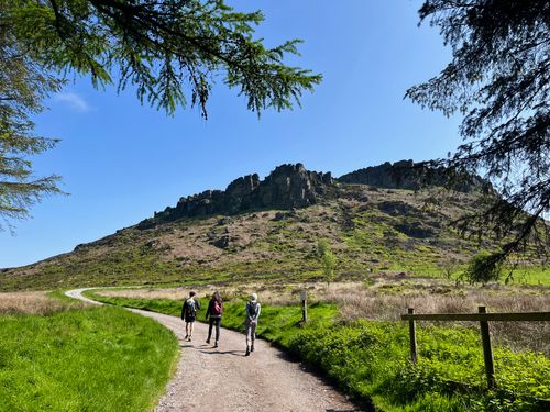 A bright, sunny day with blue skies. Three people walk ahead toward craggy rocks which reach up through the horizon.