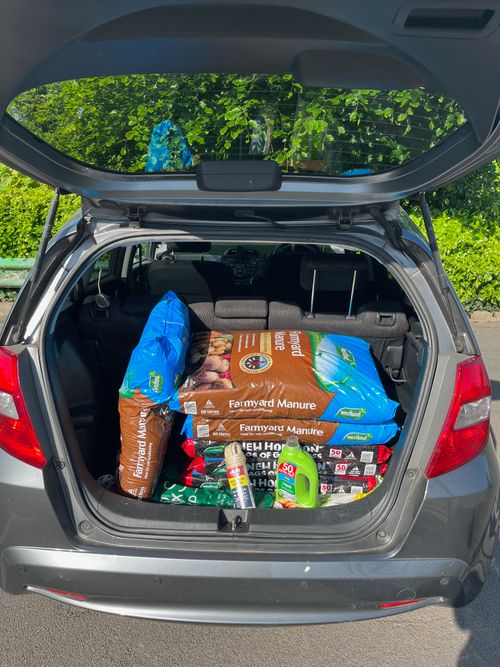 An open car boot with three bags of peat-free compost, three bags of manure, seaweed feed, and rope.